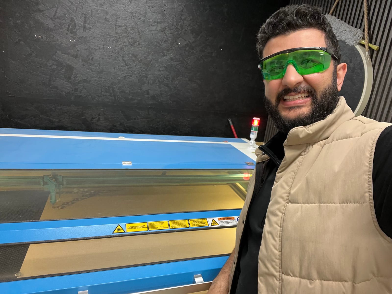 Mahmoud in green safety goggles at the laser cutter, mid-cut.